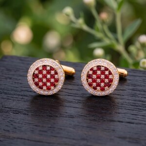 May include: A pair of round gold cufflinks featuring a central checkerboard pattern of small red stones alternating with clear stones, surrounded by a border of tiny clear pavé stones set in a circular gold frame with a standard toggle backing.