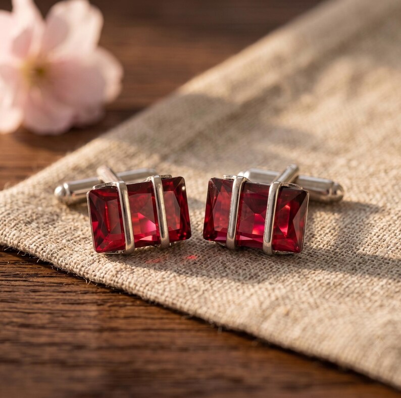 Puede incluir: Un par de gemelos rectangulares con piedras facetadas de color rojo intenso engastadas en monturas de metal plateado pulido. Cada gemelo tiene dos barras verticales plateadas que cruzan el centro de la gema roja brillante.
