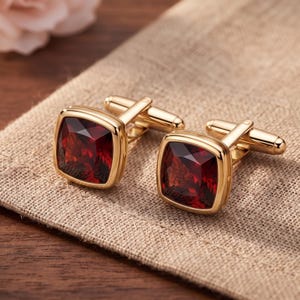 Peut inclure: Une paire de boutons de manchette dorés avec des pierres précieuses facettées rouge foncé de forme carrée, serties dans des montures en métal poli. Chaque bouton possède un mécanisme à bascule en T articulé pour la fixation sur une chemise.