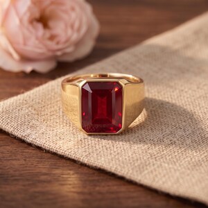 Puede incluir: Un anillo de sello de oro con una gran piedra preciosa de color rojo intenso de corte esmeralda rectangular en el centro. La banda es lisa y pulida, mostrando un diseño clásico con una piedra facetada prominente que refleja la luz.