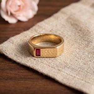 Op de afbeelding: Een goudkleurige bandring met een plat, rechthoekig bovenoppervlak waarin een enkele, kleine, verticaal georiënteerde, rechthoekig geslepen rode edelsteen verzonken ligt. De ring heeft een gepolijste, gladde afwerking en een solide, dikke constructie.