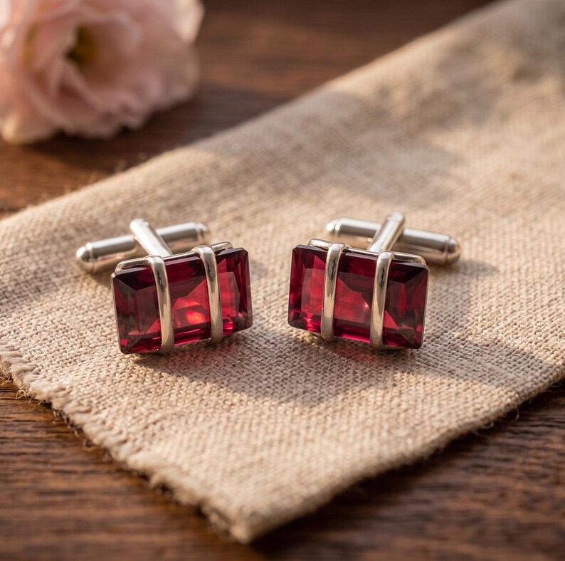 Puede incluir: Un par de gemelos rectangulares con piedras preciosas facetadas de color rojo intenso engastadas en monturas de metal plateado pulido. Cada gemelo tiene un mecanismo giratorio en forma de T en la parte posterior para su sujeci&oacute;n.