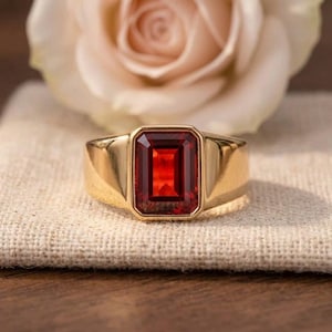 May include: A gold ring featuring a large, rectangular, emerald-cut red gemstone set in a smooth, polished metal band. The ring is positioned in front of a soft, pale pink rose, highlighting the vibrant contrast between the deep red stone and the gold.