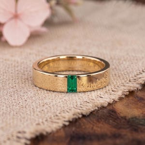 Peut inclure: Une bague en or poli avec une pierre précieuse verte taille émeraude, sertie verticalement au centre. La bague présente une surface métallique lisse et réfléchissante ainsi qu'un profil plat, offrant un design épuré avec une pierre verte.