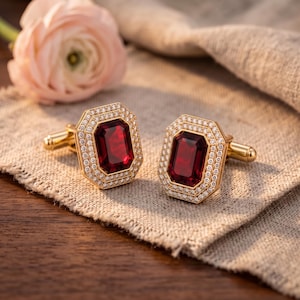 May include: A pair of gold cufflinks featuring a large, rectangular, deep red gemstone at the centre, surrounded by a double halo of small, clear, sparkling stones. The cufflinks have a polished gold finish and a standard toggle backing mechanism.