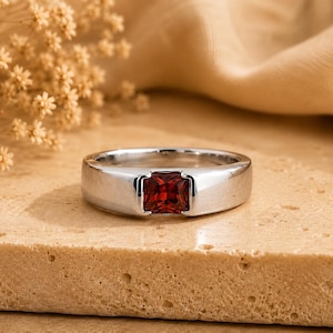 Puede incluir: Un anillo de metal plateado con una piedra preciosa de color rojo intenso de corte cuadrado, engastada de forma segura en el centro de la banda. La banda tiene un acabado liso y pulido con un ancho uniforme, resaltando el contraste del color.