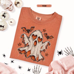 May include: A yam-colored t-shirt featuring a graphic of a ghost with heart-shaped eyes and mouth, surrounded by bats and stars. The ghost design has a distressed, frayed-edge appearance. The shirt has a crew neckline and a Comfort Colors label.