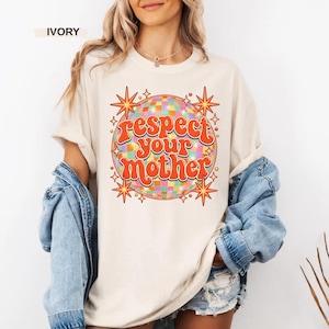May include: An ivory t-shirt featuring a graphic of a multicolored disco ball surrounded by orange starbursts. The text "respect your mother" is printed in a bold, orange, retro-style font across the center of the disco ball design.