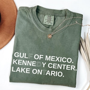 May include: An olive green short-sleeve t-shirt with a crew neckline and a Comfort Colors brand label. The shirt features white printed text that reads GULF OF MEXICO. KENNEDY CENTER. LAKE ONTARIO. A thin gold chain necklace with a small round pendant rests on it.