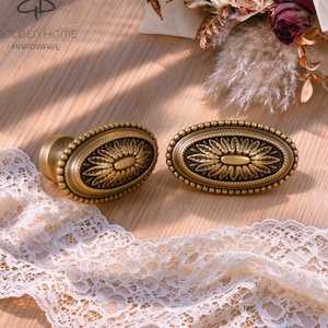 Könnte beinhalten: Zwei ovale Schrankknöpfe aus Messing mit einem zentralen geprägten Blumenmotiv, das von einem Perlenrand umgeben ist. Die Beschläge haben ein dunkles, antikes Finish mit Highlights auf den erhabenen Flächen. Der Text GOLDEN HOME HARDWARE ist sichtbar.