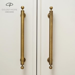 May include: Two vertical cylindrical cabinet pulls with a brushed antique brass finish. Each handle features a long, textured rod with a spherical knob at the top and bottom, mounted onto a white surface. The text GOLDEN HOME HARDWARE is visible.