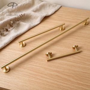 May include: Two brushed gold cabinet pulls with a cylindrical bar design and circular mounting bases. The hardware features a smooth metallic finish and a simple, linear structure. The brand name GOLDEN HOME HARDWARE is visible in the upper left corner.