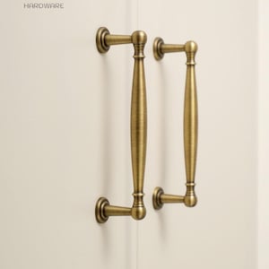 Könnte beinhalten: Zwei antike Messing-Schrankgriffe mit zylindrischem, konischem Design und abgerundeten Montagebasen. Die Beschläge sind vertikal auf einer cremefarbenen Oberfläche montiert. Das Logo von Golden Home Hardware ist oben links zu sehen.
