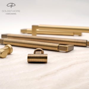 May include: A collection of brushed brass cabinet hardware including long bar pulls and T-shaped knobs. The metal pieces feature a matte finish and rectangular mounting bases. The logo for Golden Home Hardware is visible in the upper left corner.