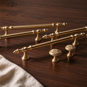 May include: A collection of gold-toned metal cabinet hardware, including three long, slender bar pulls with decorative finials and two round, floral-patterned knobs. The hardware features a smooth, brushed finish and a consistent metallic gold colour.