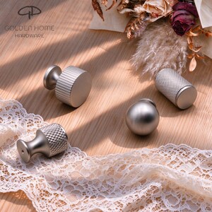 Peut inclure: Quatre boutons de placard en métal argenté aux textures variées, dont deux avec une prise moletée à motif diamant, un avec des rainures verticales et un bouton sphérique lisse, avec le logo Golden Home Hardware dans le coin.