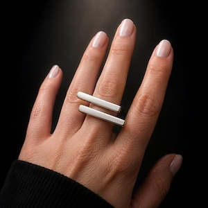 Op de afbeelding: Een zilverkleurige ring met twee parallelle horizontale staven die over de vinger lopen. Het metaal heeft een gladde, gepolijste afwerking met strakke, rechte randen, wat een geometrisch ontwerp vormt dat plat op de vinger ligt.