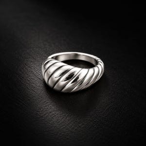 May include: A polished silver-toned ring featuring a thick, rounded band with a series of diagonal, grooved ridges that wrap around the exterior surface, creating a textured, croissant-like appearance with a high-shine metallic finish.