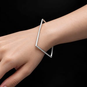 Op de afbeelding: Een zilverkleurige armband met een stijf, geometrisch rechthoekig frame. De armband is opgebouwd uit dunne, gladde metalen staven die een scherpe, hoekige vorm vormen en de pols omsluiten met een strakke en gepolijste metalen afwerking.