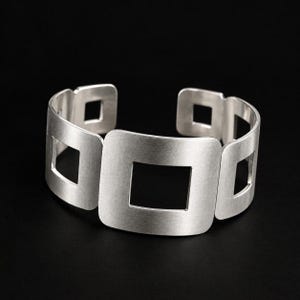 May include: A silver-toned cuff bracelet featuring a series of connected square-shaped links with open centers. The metal has a matte, brushed finish and a wide, rigid band design that forms an open circle for the wrist.