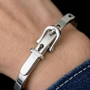May include: A polished silver-tone metal bangle bracelet designed to resemble a miniature belt. The bracelet features a functional-style buckle mechanism with a prong, a rectangular frame, and a smooth, reflective surface that wraps around the wrist.