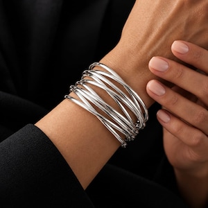 Può includere: Un bracciale a fascia color argento composto da diversi fili metallici sottili e lucidi che si sovrappongono e si intrecciano per creare un design ampio e stratificato, che avvolge il polso con una finitura metallica liscia e riflettente.