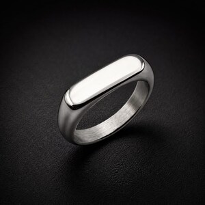Op de afbeelding: Een ring van gepolijst zilverkleurig metaal met een gladde, langwerpige rechthoekige bovenplaat met afgeronde randen. De ring heeft een solide, dikke constructie met een reflecterende afwerking die het licht opvangt voor een strak uiterlijk.