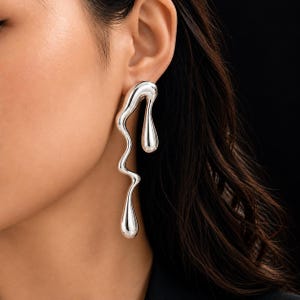 Peut inclure: Une longue boucle d'oreille argentée au design fluide évoquant du métal en fusion. La structure verticale ondulée se termine par deux formes de gouttes lisses et arrondies, créant un aspect rappelant un liquide en mouvement.