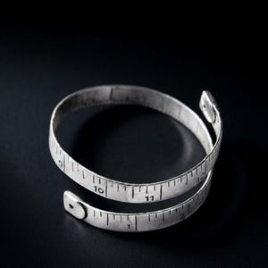 Peut inclure: Un bracelet enroulable en métal argenté conçu pour ressembler à un ruban à mesurer flexible. La bande présente des marques et des chiffres noirs gravés allant de 1 à 12, avec une finition plate et lisse et des extrémités arrondies en spirale.