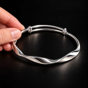 May include: A polished silver-tone bangle bracelet featuring a continuous twisted, wavy design. The bracelet has an adjustable sliding mechanism with two parallel metal bars at the top, allowing the diameter to expand and contract for a secure fit.