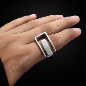 May include: A polished silver-toned ring featuring a geometric design with an open rectangular frame and a central vertical bar element. The metal surface is smooth and reflective, showcasing a clean, structural aesthetic that wraps around the finger.