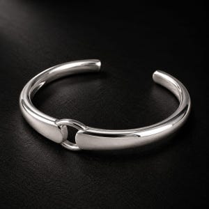 May include: A polished silver cuff bracelet featuring a smooth, rounded band that tapers toward the center. The design includes a central circular ring connector that joins the two ends of the metal band, creating an open-ended, circular silhouette.