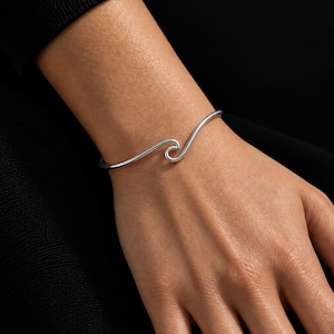 May include: A silver-toned metal cuff bracelet featuring a continuous, smooth, and polished wave-shaped design that curves into a central spiral element. The bracelet has a sleek, minimalist, and open-ended structure that rests on the wrist.