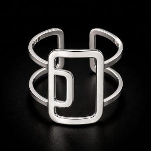 Könnte beinhalten: Ein offener Ring aus poliertem silberfarbenem Metall mit einem zentralen rechteckigen Rahmendesign mit abgerundeten Ecken und einem kleineren, innenliegenden rechteckigen Ausschnitt. Die Ringschiene besteht aus zwei parallelen Drähten.