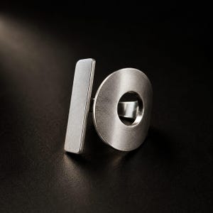 May include: A silver-toned metal ring featuring a brushed finish. The design consists of a vertical rectangular bar positioned next to a circular disc with a central cutout, creating a geometric composition with a smooth, matte metallic texture.
