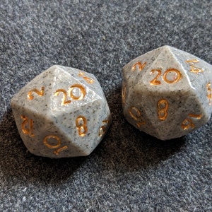 May include: Two grey 20-sided dice with gold numbers. The dice are speckled with small white flecks.