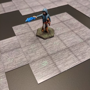 May include: Miniature fantasy figure of a hooded character in a blue jacket and tan pants, holding a blue crystal. The figure stands on a hexagonal base, placed on a gray, brick-patterned game board.