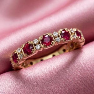 Puede incluir: Un anillo de banda de oro con una serie de rubíes rojos redondos y pequeños diamantes blancos transparentes alternados. Las piedras preciosas están engastadas en un patrón repetitivo dentro de un marco de metal dorado texturizado.