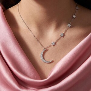 May include: A silver-toned necklace featuring a crescent moon pendant and a series of small star charms along the chain. The moon and stars are encrusted with small, clear, sparkling stones that catch the light, creating a bright, reflective finish.