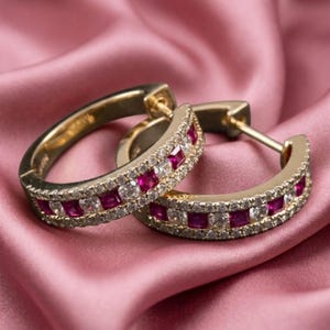May include: A pair of gold hoop earrings featuring a central row of square-cut vibrant red rubies flanked by two rows of small, clear, round-cut white diamonds. The earrings have a hinged post closure and a polished metallic finish.