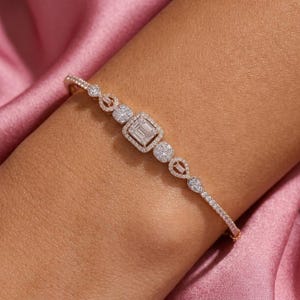 Op de afbeelding: Een roségouden armband met een centrale rechthoekige diamant in emerald-slijpvorm, omringd door een halo van kleinere ronde diamanten, geflankeerd door extra ronde en baguette-geslepen diamanten op een fijne metalen ketting.