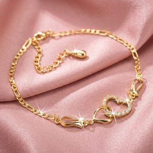 May include: A gold-tone chain bracelet featuring a central heart-shaped charm encrusted with small clear stones. The bracelet includes a delicate link chain, a secondary heart-shaped metal element, and an adjustable extender chain with a small drop charm.