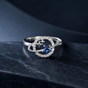 May include: A silver-toned ring featuring a circular design resembling a planet with a ring around it. The circular frame is encrusted with small clear stones, while the center contains a blue star-shaped stone and two small silver star cutouts.