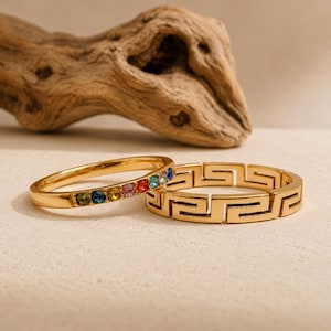 Puede incluir: Dos anillos de tono dorado. Un anillo presenta una hilera de pequeñas piedras preciosas redondas y multicolores en tonos azul, verde, amarillo, rosa y rojo. El segundo anillo muestra un patrón geométrico griego continuo y repetitivo.