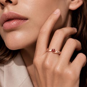 Puede incluir: Un anillo de oro rosa con una piedra preciosa redonda de color rojo intenso engastada en el centro. La banda tiene un acabado liso y pulido e incluye el nombre Enna grabado en el lateral. El diseño es de estilo bypass donde los extremos se unen.