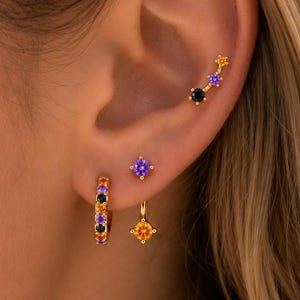 May include: A set of gold-toned earrings featuring a small hoop with alternating black and purple stones, a single purple stud, a gold stud with an orange stone, and a curved ear climber with black, purple, and orange stones in a star-like setting.