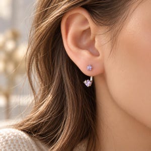 Puede incluir: Un pendiente de plata con un pequeño perno de estrella morada en el lóbulo y una barra plateada curva que se extiende detrás, terminando en una gema morada de corte cuadrado con un pequeño detalle de estrella plateada al lado.