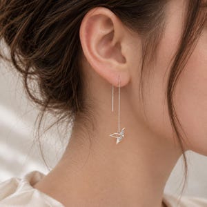 May include: A silver-toned threader earring featuring a delicate, geometric origami crane pendant. The earring consists of a thin metal chain that passes through the earlobe, ending in a small, angular bird shape with fine, clean lines and a polished finish.
