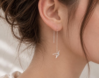 May include: A silver-toned threader earring featuring a delicate, geometric origami crane pendant. The earring consists of a thin metal chain that passes through the earlobe, ending in a small, angular bird shape with fine, clean lines and a polished finish.