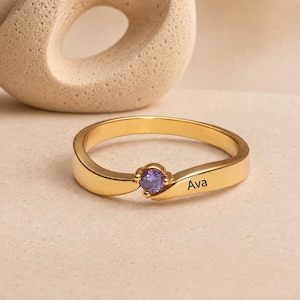 Puede incluir: Un anillo de tono dorado con una banda lisa y curva que presenta una piedra preciosa redonda de color morado engastada en el centro. El nombre Ava está grabado en letras negras en el lateral de la banda, con un acabado pulido y diseño abierto.